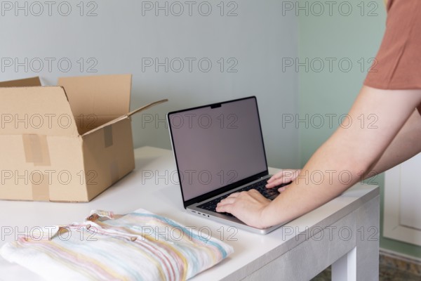 A person uses a laptop to organize and prepare clothing for online sale. The scene includes a striped shirt and a cardboard box, representing decluttering and entrepreneurship