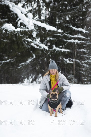 A woman in a winter coat kneels with a dog in a snow-covered forest. The serene winter landscape adds charm to this peaceful pet outing in nature