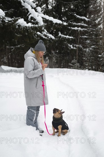 A woman in a warm coat and hat walks her dog on a snowy trail. The forest around is covered in snow, emphasizing the serene winter atmosphere