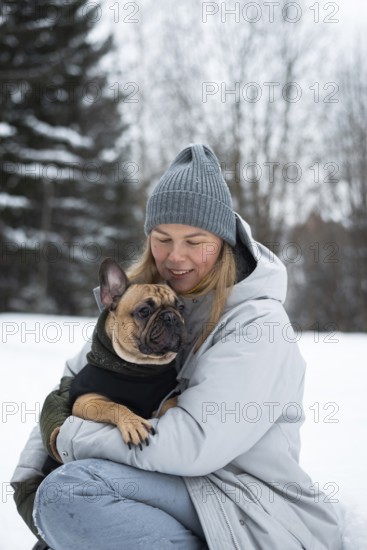 A woman in winter attire lovingly holds her French Bulldog in a snowy landscape. The scene captures the warmth and companionship between them amidst the cold surroundings