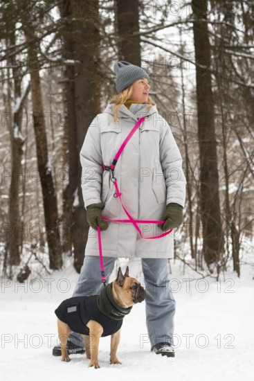 A woman in a winter coat and hat walks a French Bulldog through a snowy forest. The dog wears a warm jacket, and a bright pink leash adds a pop of color