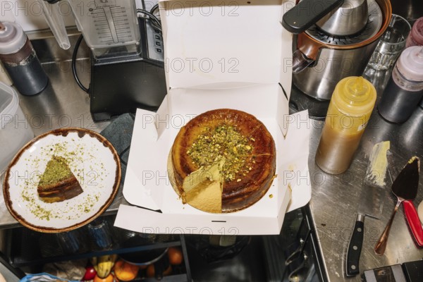 A freshly baked cheesecake topped with crushed pistachios sits in an open box, with a slice plated nearby. The scene captures a coffee shop kitchen with utensils and ingredients