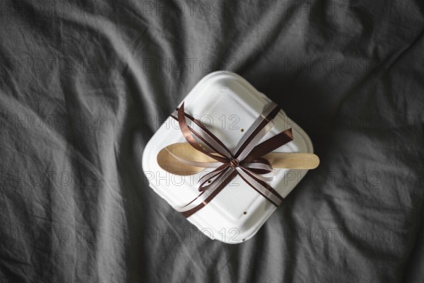 Top view of an elegantly wrapped bento birthday cake, set in a white takeout box with a brown ribbon and wooden cutlery, against a soft grey fabric backdrop