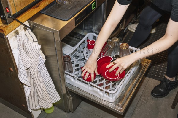 A person loads a dishwasher with red coffee cups and glass bottles, capturing the behind-the-scenes operations in a specialty coffee shop or cafe environment