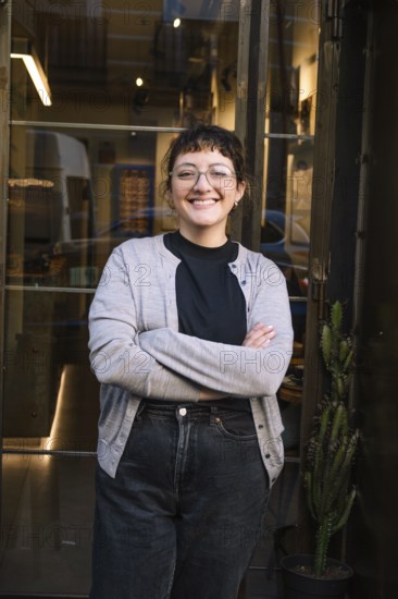 Friendly barista smiling confidently outside a cozy specialty coffee shop. Glass door entrance with warm ambiance inside, inviting customers to enjoy a unique coffee experience