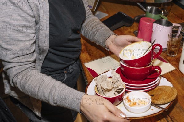 A person collects red cups with coffee grounds and plates on a wooden table in a coffee shop. This scene captures the busy atmosphere of a specialty coffee shop