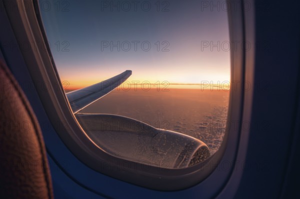 View through an airplane window catching the glowing sunset skyline above the clouds, with the plane's wing prominently displayed against the horizon