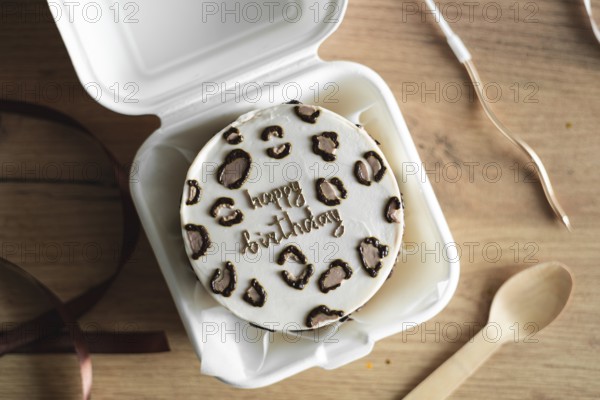 Top view of a stylish Bento birthday cake decorated with heart-shaped toppings, presented in a white takeout box with a wooden spoon alongside, set on a rustic wooden table