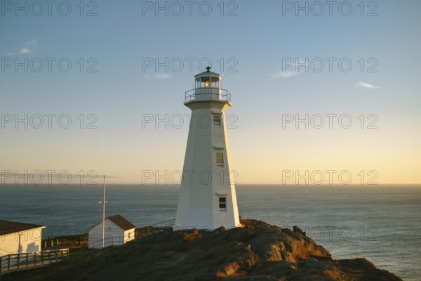 A warm sunset casts a golden glow over the white structure of Cape Spear Lighthouse, perceived as the most easterly point in North America, located in St. John's, Newfoundland & Labrador, Canada