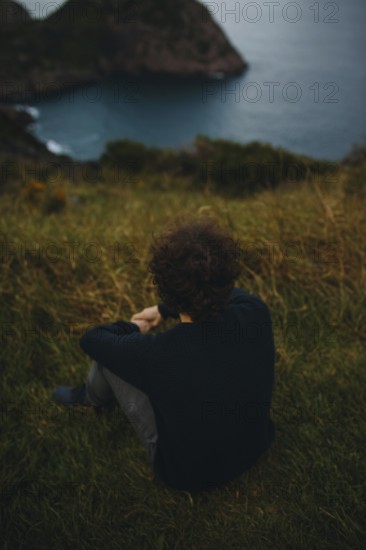 Back view of an unrecognizable man seated on grassy terrain, gazing towards a distant sea with cliffs under a moody sky in Signal Hill in St. John's, Canada