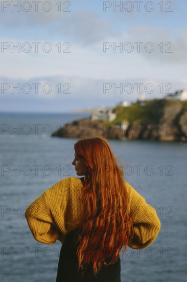 Back view of an unrecognizable woman with long red hair, standing at Cape Spear Lighthouse, Newfoundland & Labrador, Canada, overlooking the ocean