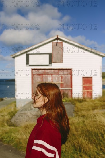 A young thoughtful woman with long red hair and eyes closed, wearing a red jacket, stands contemplatively in front of a white barn with red doors in Flatrock, Newfoundland & Labrador, with the ocean in the background under a blue sky