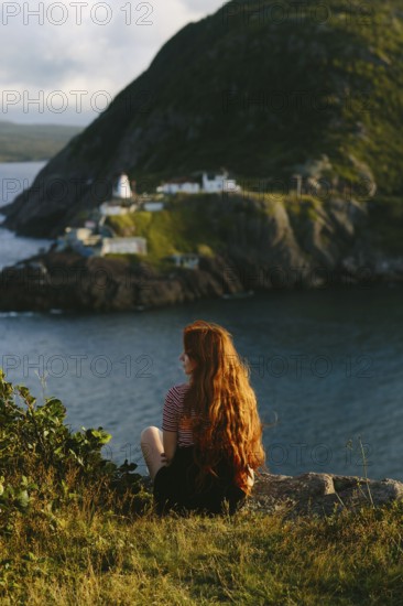 A woman with long red hair sits contemplatively overlooking the dramatic landscape of Cape Spear Lighthouse, Newfoundland & Labrador, Canada. The image captures a serene moment with a soft golden hour light illuminating the sea and rugged cliffs