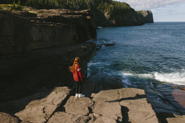Back view of a young unrecognizable redhead woman standing on the rocky shoreline of Flatrock, Newfoundland, looking out at the powerful ocean waves. She wears a red jacket and white shoes, with her long hair catching the breeze