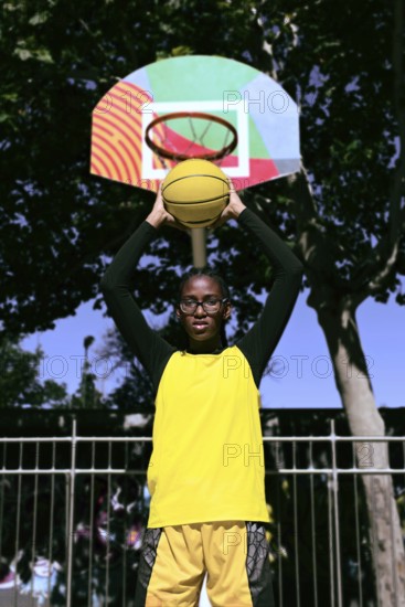 A black basketball player stands ready to shoot, holding a yellow basketball toward a vibrant, patterned basket hoop. The scene captures the spirit of urban sports in a park setting