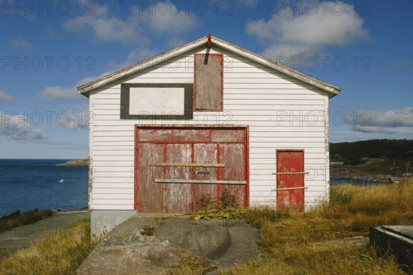 A weathered white and red storage building in Flatrock, Newfoundland & Labrador, Canada. The structure, overlooking the Atlantic Ocean, has a rustic charm with faded paint and wooden details against a backdrop of blue skies and scattered clouds