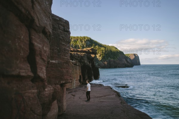 A young man standing on a rocky cliff overlooking the ocean at Flatrock, Newfoundland and Labrador, Canada. The scene captures serene natural beauty and a moment of solitude