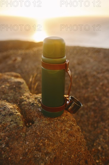 A thermos sits on a rugged rock with the warm sunset illuminating the ocean backdrop in Cape Spear Lighthouse, Newfoundland. The serene scene depicts an early morning outdoor adventure mood