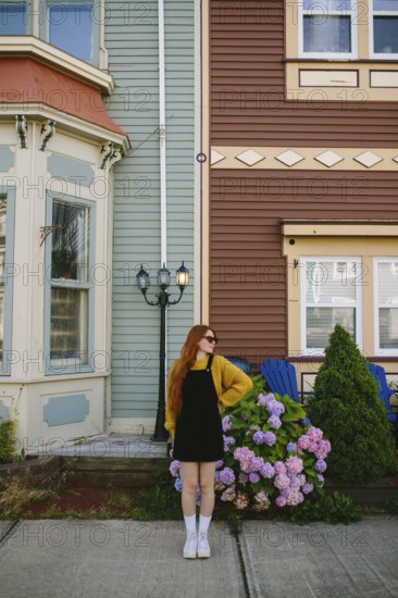 A young woman with red hair stands beside classic architecture in St. John's, Newfoundland & Labrador, Canada. Dressed in a stylish black dress and a vibrant yellow cardigan, she enjoys the charming surroundings