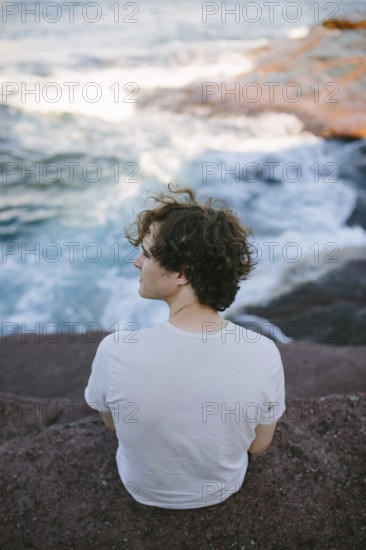 Back view of a young man at Flatrock looking out at the turbulent ocean waves and rocky coastline, reflecting a moment of solitude and nature's beauty