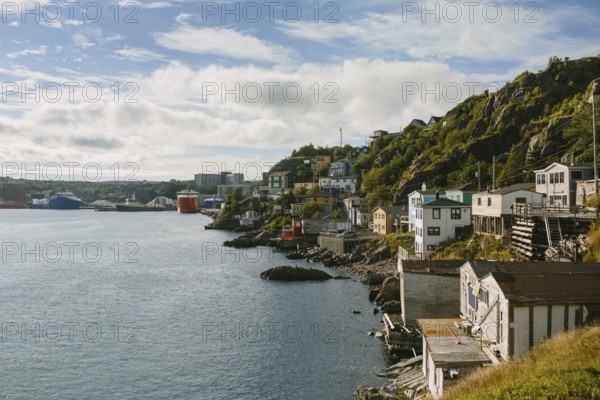 Coastal cityscape of St. John's, Newfoundland & Labrador, showcasing quaint, multicolored houses perched on rocky hillsides along the waterfront, under a partly cloudy sky