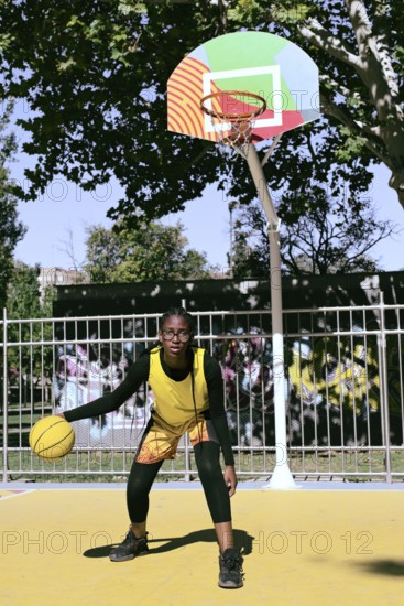 A woman athlete in a yellow jersey dribbles a basketball on an outdoor court with a colorful hoop. The scene is set in a sunlit park surrounded by trees