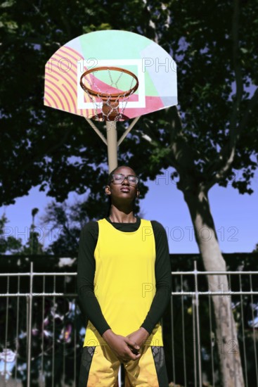 A woman basketball player in a yellow jersey stands confidently beneath a colorful hoop. The scene captures the spirit of sport and determination in an outdoor setting
