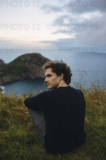 A young male sits serenely overlooking the calm waters from Signal Hill, St. John's. His relaxed pose contrasts with the wild, natural setting around him, capturing a moment of peace