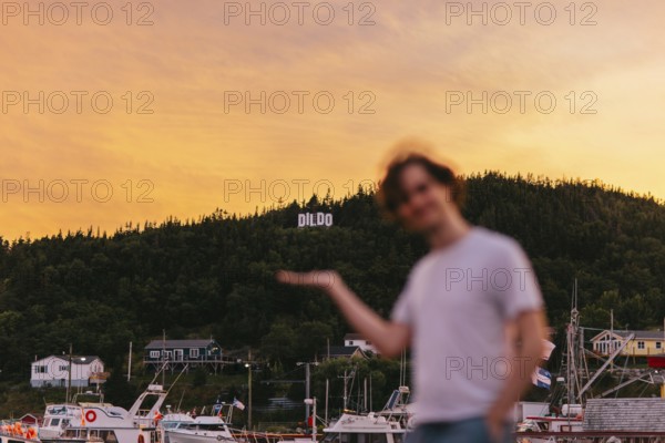 A young blurred Caucasian man playfully gestures towards the town sign DILDO in Dildo, Newfoundland & Labrador, Canada. The blurry sunset backdrop highlights lively harbor activity