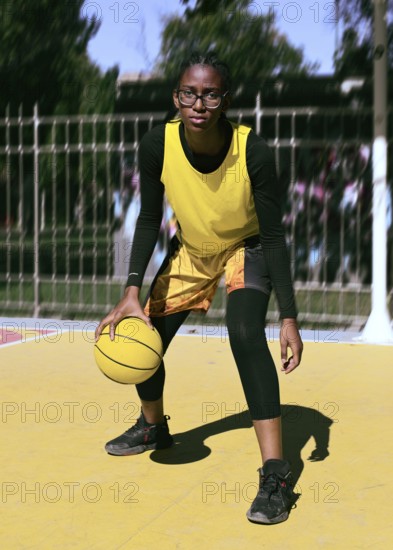A focused woman basketball player dribbles a ball on a vibrant outdoor court. Dressed in a yellow jersey, they practice their skills under bright sunlight, embodying athleticism