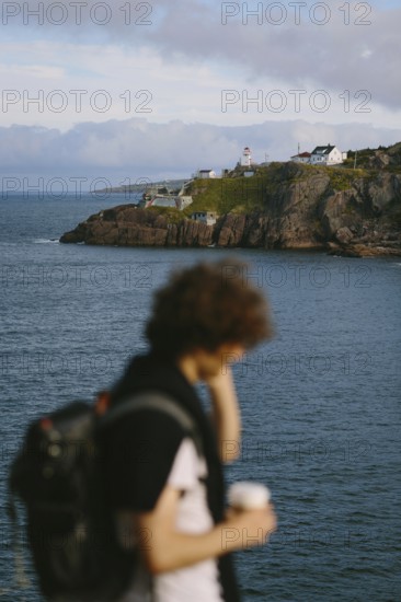 Unrecognizable man with curly hair, sipping coffee while overlooking the rugged coastline and the Fort Amherst lighthouse at Signal Hill, St. John's, Newfoundland & Labrador, Canada