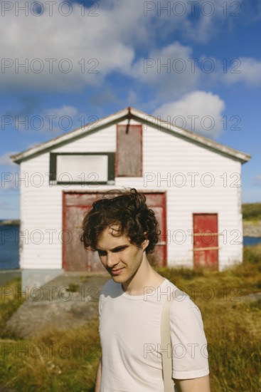A young man with curly hair stands outdoors, smiling subtly near an old white building with red trim in Flatrock. His casual appearance and the clear, sunny backdrop create a peaceful, scenic image