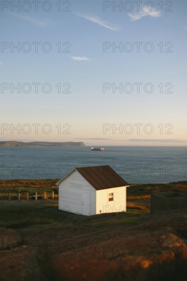 A lone white shed stands on the rugged terrain of St. John's, Newfoundland & Labrador, Canada, bathed in the warm glow of the setting sun. The distant hills and a serene sea that stretches to the horizon complete this peaceful seascape