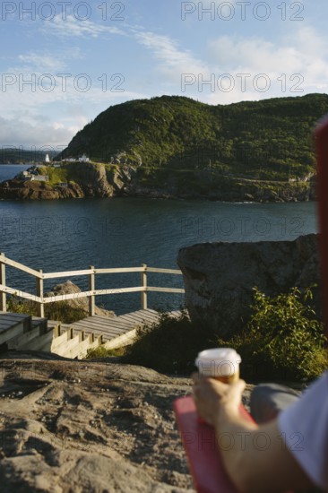 Cropped unrecognizable person sitting on rocks at Signal Hill, holding a coffee cup with a scenic overlook of the Fort Amherst in the rugged coastline and green hills of St. John's in Newfoundland & Labrador, Canada