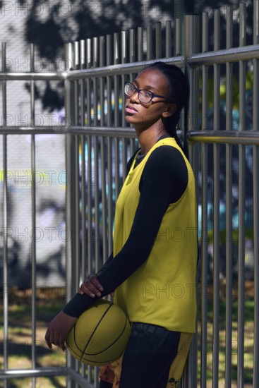 A woman basketball player in a yellow jersey and glasses leans against a metal fence, holding a basketball in an urban outdoor setting under sunlight