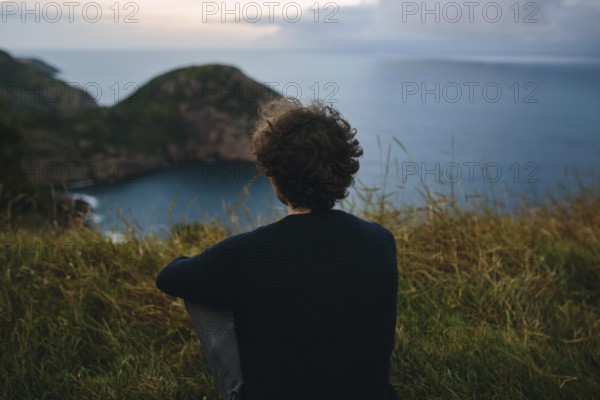Back view of unrecognizable person sitting on grassy hill, overlooking a secluded coastal inlet beneath a cloudy sky at Signal Hill in St. John's, Newfoundland & Labrador, Canada