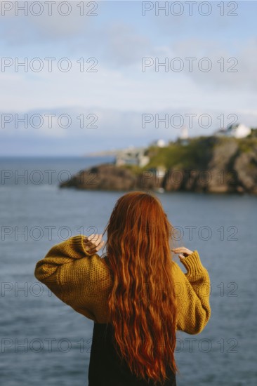 Back view of unrecognizable woman with long red hair, wearing a yellow sweater, overlooking a picturesque ocean cliff in Signal Hill, St. John's, Newfoundland & Labrador, Canada