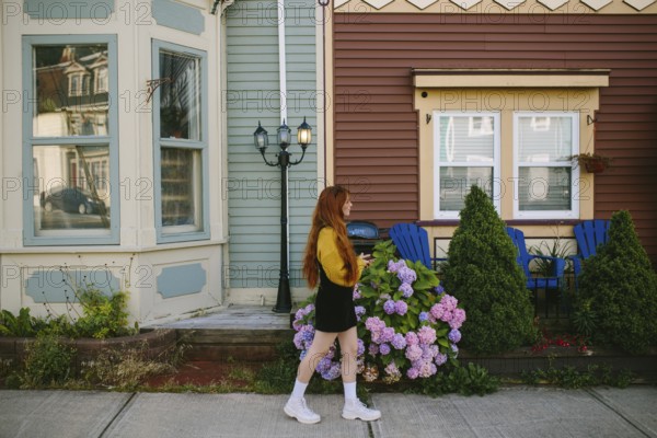 A young woman walks past charming houses with vividly painted facades in St John's, Newfoundland & Labrador. She is enjoying the vibrant street adorned with colorful flowers and quaint architectural details