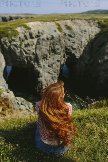 Back view of an unrecognizable redhead woman sitting on a grassy edge, overlooking a rugged coastal landscape in Dungeon Provincial Park, Newfoundland & Labrador, Canada