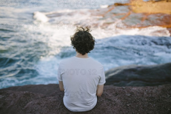 Back view of an unrecognizable person sitting on a rock by the sea, enjoying the serene view at Flatrock, Newfoundland & Labrador, Canada