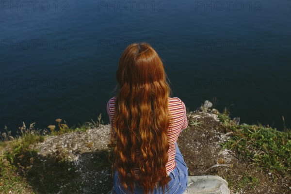 Back view of an unrecognizable redhead female sitting and looking out over the ocean from a cliff. She is wearing a striped red-and-white T-shirt and blue jeans, enjoying a serene moment at Dungeon Provincial Park, Newfoundland & Labrador, Canada