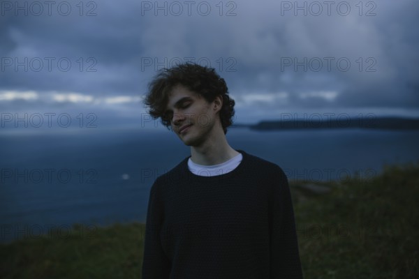 A thoughtful young man with eyes closed stands by the ocean in Signal Hill in St. John's, Newfoundland & Labrador, Canada. And he is lost in contemplation against a dramatic, cloudy seascape
