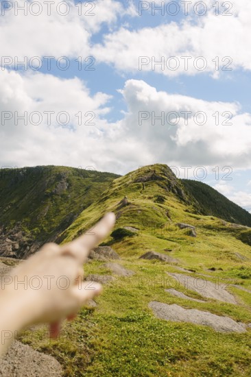 Cropped shot of unrecognizable hand pointing towards a lush green hill under a partly cloudy sky in Middle Cove, Newfoundland & Labrador, Canada. The focus is on the vibrant landscape and dramatic clouds above
