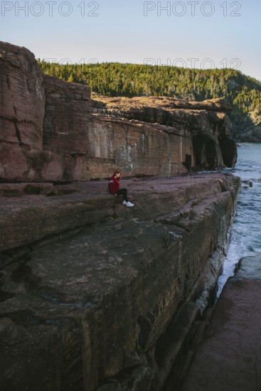 A woman in a red jacket sits pensively on a large rock cliffside, overlooking the ocean at Flatrock, Newfoundland & Labrador, Canada. The scene is calm and serene, set against a backdrop of forested hills and clear skies