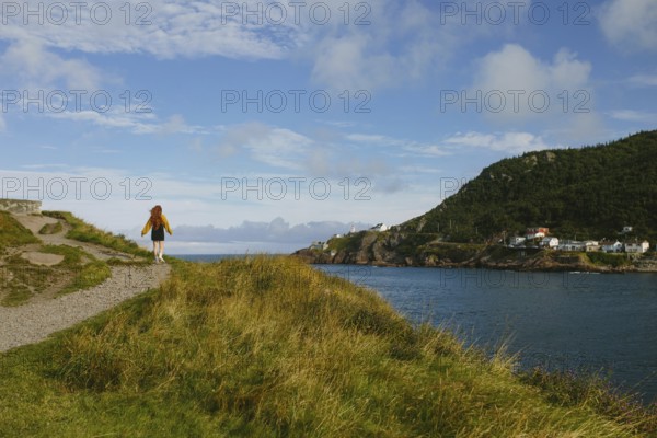 Back view of unrecognizable woman enjoying a serene walk along a coastal trail in Signal Hill, St John's, overlooking the rugged cliffs and calming waters of Newfoundland & Labrador. The clear blue sky and lush green grass enhance the tranquil scene