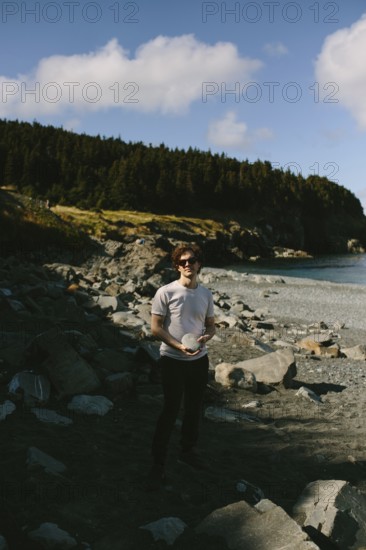 A young man stands on the rocky shores of Middle Cove in Newfoundland & Labrador, Canada. He is casually dressed, holding a hat and surrounded by a rugged coastal landscape