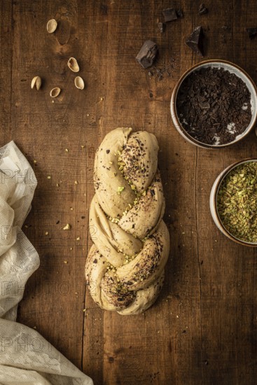 Top view of a freshly baked Bakbah bread adorned with chocolate shavings and pistachio bits, placed on a rustic wooden table surrounded by ingredients