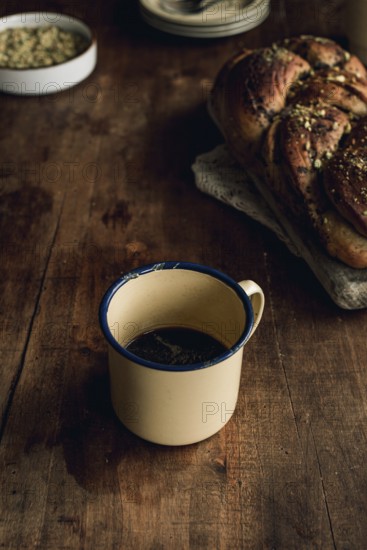 Top view of a freshly baked Bakbah bread garnished with pistachio, alongside a vintage tin cup filled with black coffee, presented on a rustic wooden table
