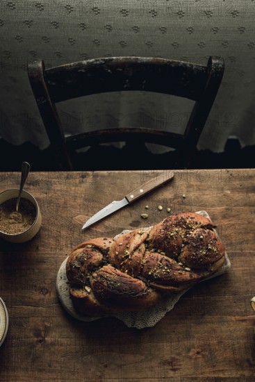 Top view of a freshly baked chocolate and pistachio babka bread, placed on a rustic wooden table beside a bowl of sugar and a knife. The image captures the bread's rich texture and the cozy, homely background hints at a traditional baking setting
