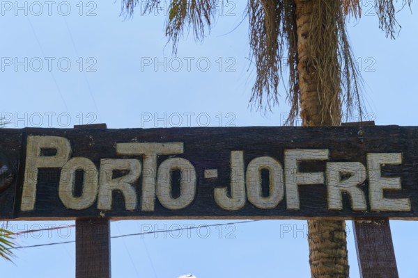 A wooden sign with the inscription 'Porto-Jofre' hangs under palm trees against a blue sky, Porto Jofre, Pantanal, Mato Grosso, Brazil